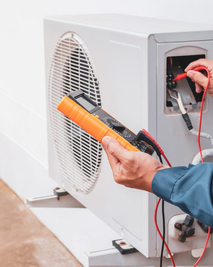 Hands delicately wield a voltmeter to perform a thorough maintenance check on an air conditioner unit while the voltmeter's probes make contact with strategic points on the unit's wiring